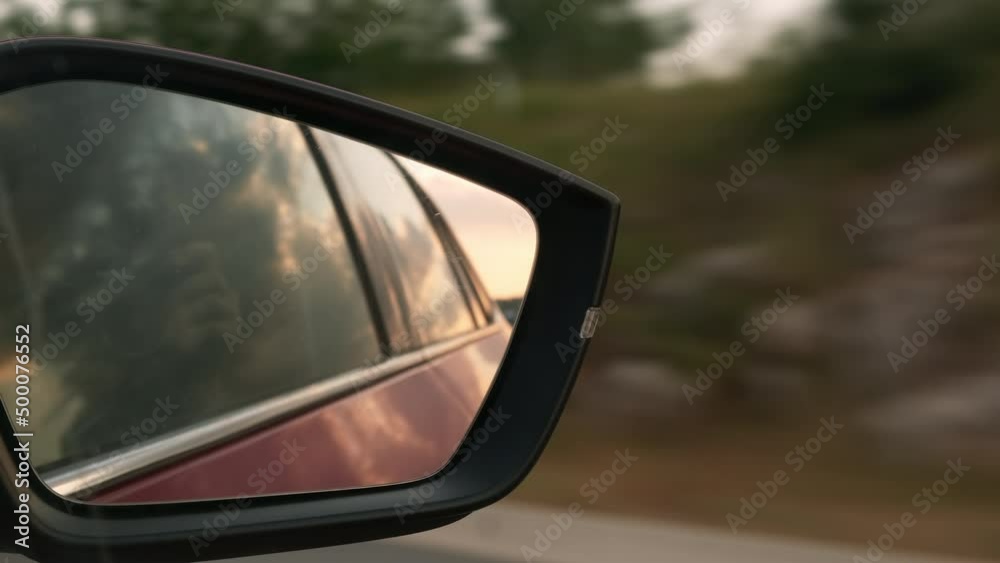 Reflection of doors in side-mirror of car driving past rows of trees growing on roadside. Cloudy sky reflects in window glass of speeding car closeup Reflection of doors in side-mirror of car driving past rows of trees growing on roadside. Cloudy sky reflects in window glass of speeding car closeup