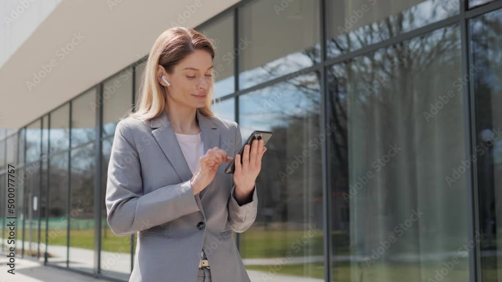 Handsome young businesswoman using a mobile phone and wireless headphones. Sending text messages while standing near a modern office building. Social network Applications Smartphone Business People. Handsome young businesswoman using a mobile phone and wireless headphones. Sending text messages while standing near a modern office building. Social network Applications Smartphone Business People.