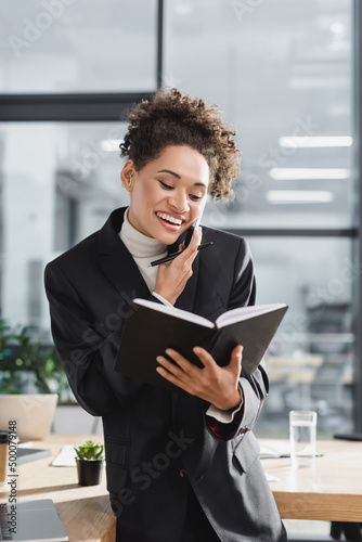 Positive african american businesswoman holding notebook and talking on smartphone in office.