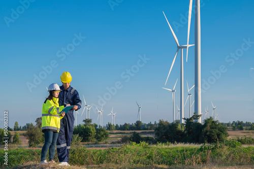 Two engineers working and holding the report at wind turbine farm Power Generator Station on mountain,Thailand people,Technician man and woman discuss about work