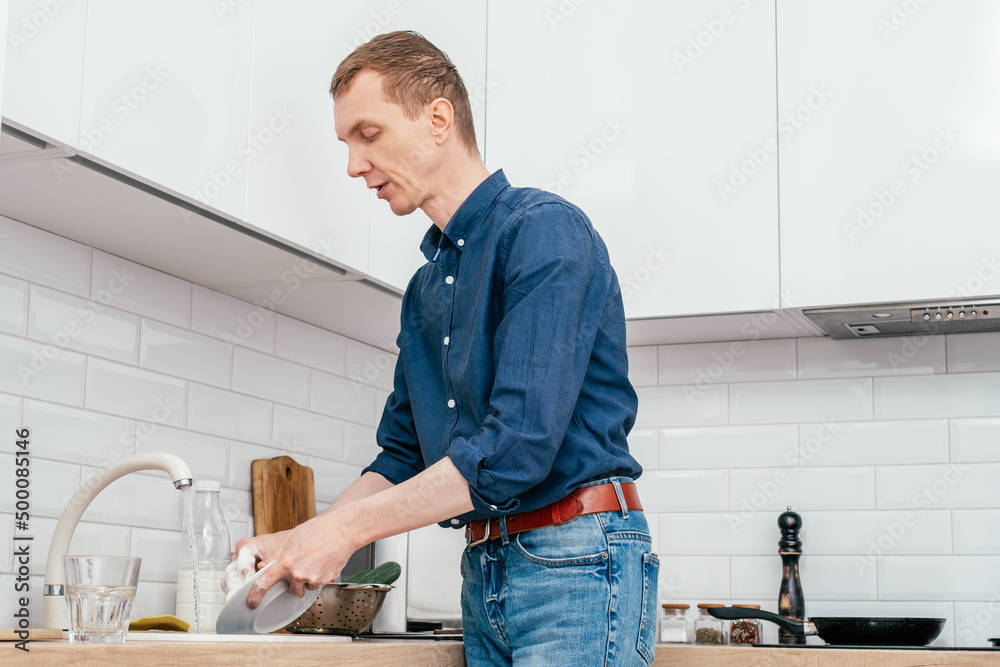 Middle-aged man with short ginger hair in blue shirt washing up white plate in beige marble kitchen sink under running water from faucet standing in white modern kitchen. Housework, cleaning concept. Middle-aged man with short ginger hair in blue shirt washing up white plate in beige marble kitchen sink under running water from faucet standing in white modern kitchen. Housework, cleaning concept.