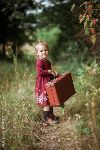 Portrait of a charming little girl in burgundy boho dress holding her old suitcase in the forest at summer time, retro style © Olena Zelena
