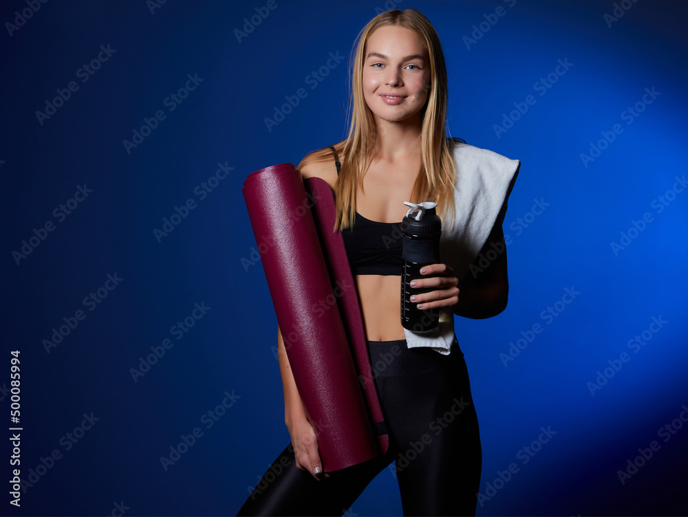 Smiling fitness woman in top and leggings takes break after training, holds bottle of water, wipes sweat with towel, being energetic runner or jogger, feels thirsty.