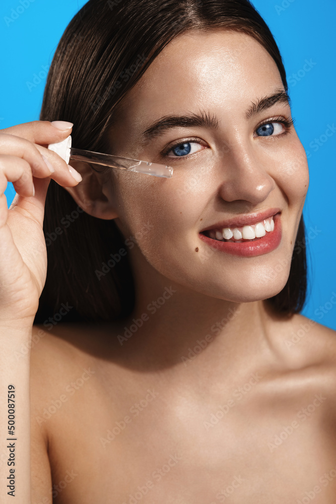 Beautiful girl applying vitamin serum on face. Young woman with long hair holding pipette of skincare product, holding dropper with cosmetic serum, isolated on studio background Beautiful girl applying vitamin serum on face. Young woman with long hair holding pipette of skincare product, holding dropper with cosmetic serum, isolated on studio background