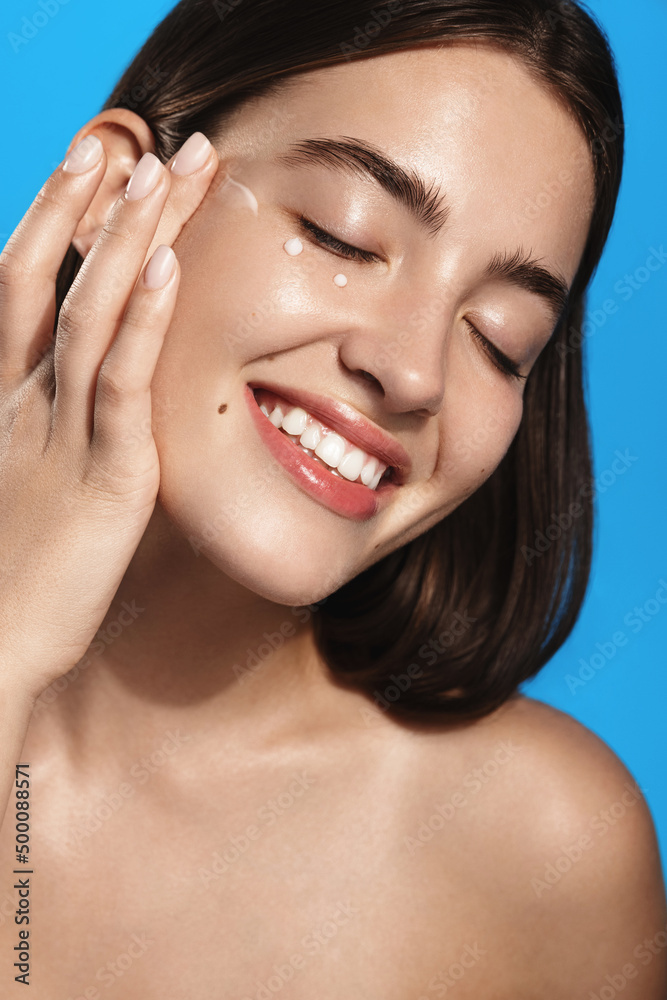 Close up of girl applies lotion, facial skin care cream product on face and smiles from pleasure, nourish face with spa cosmetic product, stands over blue background Close up of girl applies lotion, facial skin care cream product on face and smiles from pleasure, nourish face with spa cosmetic product, stands over blue background