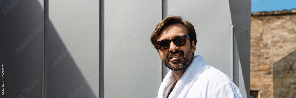 Smiling mid aged bearded man in bathrobe drinking coffee in the morning sitting on a balcony Smiling mid aged bearded man in bathrobe drinking coffee in the morning sitting on a balcony