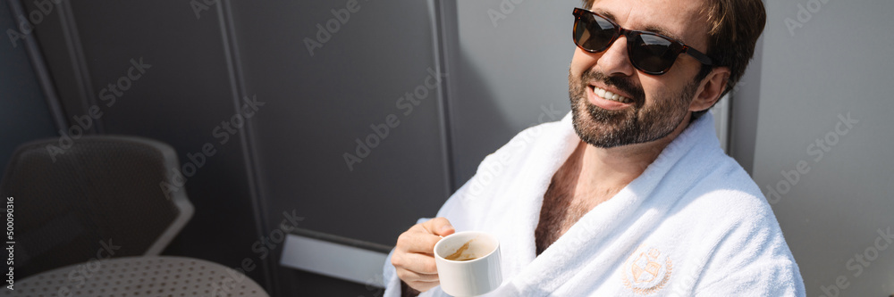 Smiling mid aged bearded man in bathrobe drinking coffee in the morning sitting on a balcony Smiling mid aged bearded man in bathrobe drinking coffee in the morning sitting on a balcony
