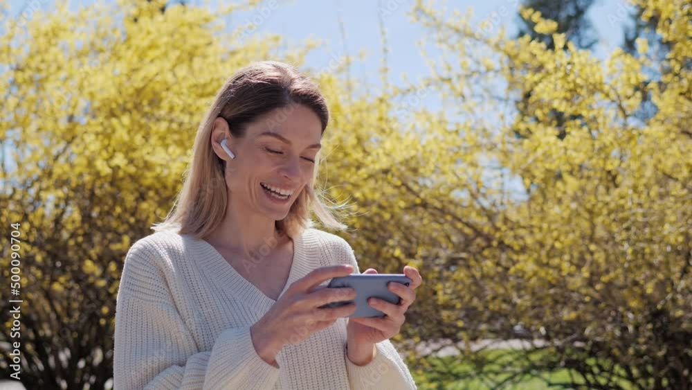 Female attractive lady in a white sweater using mobile phone woman play new game on smartphone spending free time wearing wireless headphones in the park near a tree with yellow leaves on a sunny day. Female attractive lady in a white sweater using mobile phone woman play new game on smartphone spending free time wearing wireless headphones in the park near a tree with yellow leaves on a sunny day.