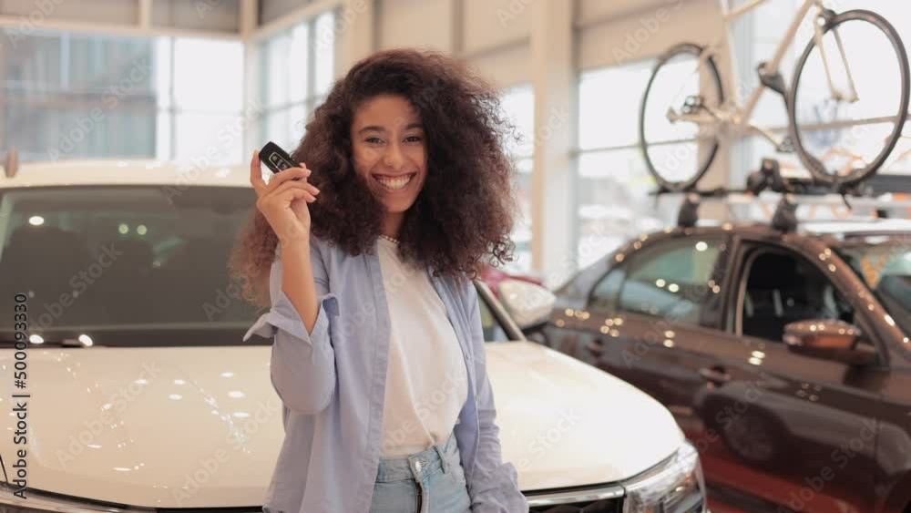 Young female client is holding keys and posing on background of new car in modern company spbi. Portrait of beautiful caucasian female looks with happy smile and holds keys in hands, poses for camera Young female client is holding keys and posing on background of new car in modern company spbi. Portrait of beautiful caucasian female looks with happy smile and holds keys in hands, poses for camera