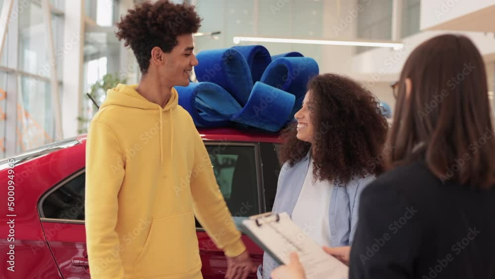 Female agent is giving keys to couple of clients during working day in modern car company spbi. Happy people buyers receive keys from seller and look with smiles, hug and stand on on background of new