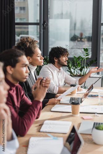 Side view of indian businessman talking near devices and blurred multiethnic colleagues during meeting in office. © LIGHTFIELD STUDIOS