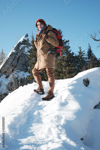 Vertical low angle shot of active young man wearing warm clothes and backpack hiking to mountain top on winter day