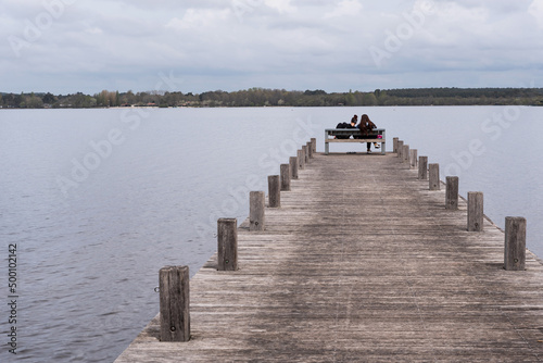 Two young women sitting on a bench on a pier at the Pond of Leon. Landes