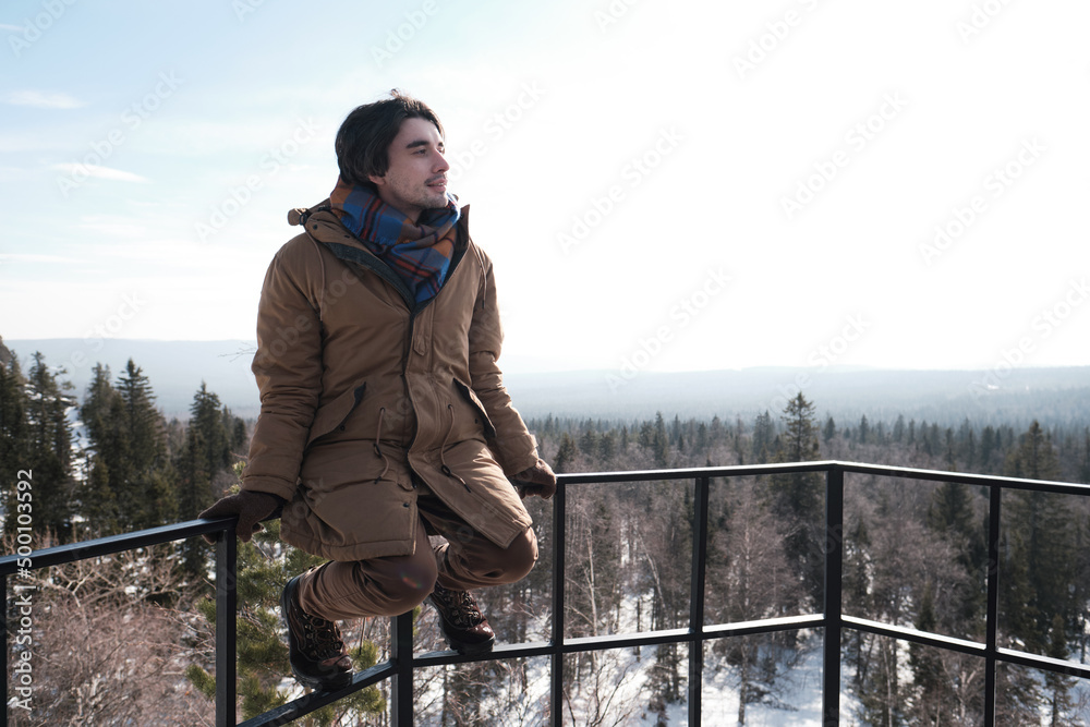 Young Caucasian man sitting relaxed on protection fence at observation deck on mountain top looking away