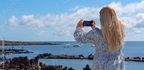 Una mujer joven rubia de espaldas sacando una foto con el teléfono móvil en la costa de Lanzarote en las Islas Canarias. Turismo veraniego.  photo