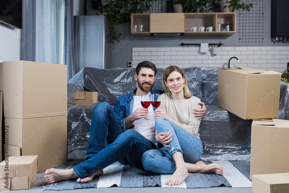 Family couple in love, man and woman sitting on the floor celebrating a new home in a new apartment, drinking wine among cardboard boxes Family couple in love, man and woman sitting on the floor celebrating a new home in a new apartment, drinking wine among cardboard boxes