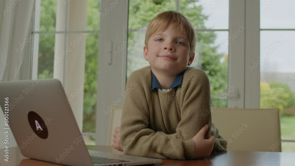 School boy smiling looking at camera sitting at the table by the window at home. Cute blonde kid is happy to learn online. School boy smiling looking at camera sitting at the table by the window at home. Cute blonde kid is happy to learn online.