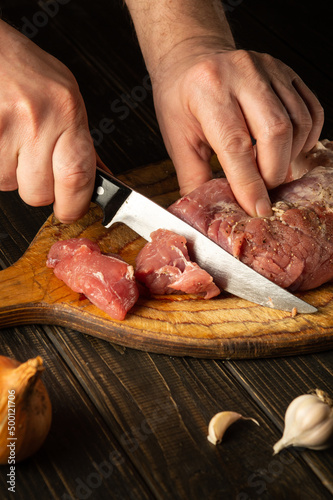 The hands of the cook cut raw beef meat on a cutting board before baking. Peasant foods