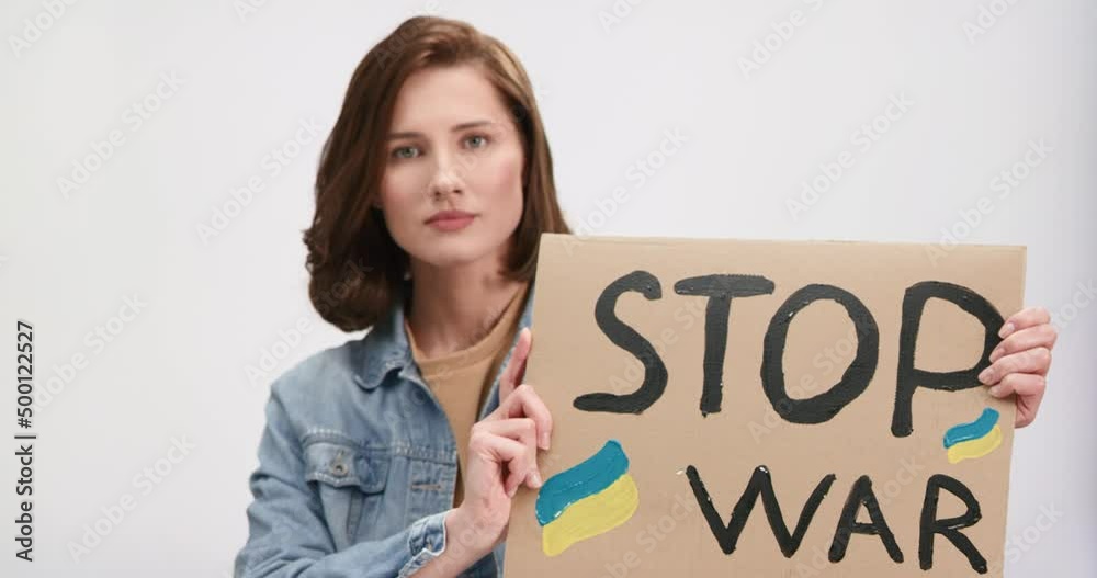 Confident caucasian woman with brown hair holding cardboard banner with Ukrainian flag and text stop war. Female activist standing in studio with white background.