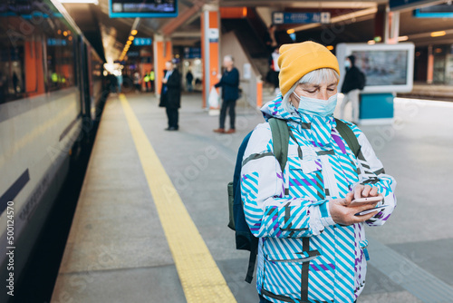 Old woman in yellow hat waiting on station platform with backpack on background train using smart phone. Railroad transport concept, Traveler © mdyn