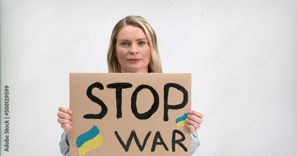 Portrait of mature blonde female in casual attire raising Stop war placard and spelling it while posing in studio. Serious woman protesting against russian invasion and violence. Portrait of mature blonde female in casual attire raising Stop war placard and spelling it while posing in studio. Serious woman protesting against russian invasion and violence.