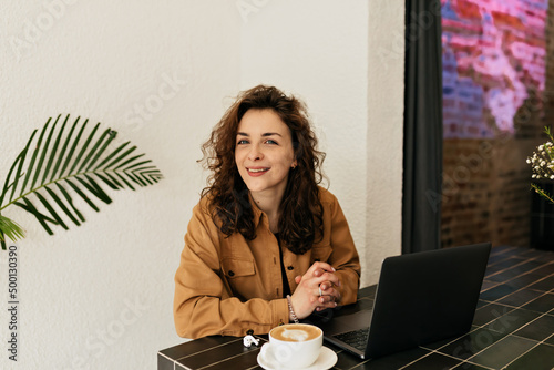 Charming girl with dark curly hair with smile looks into camera, posing at table in cafe with cappuccino and working on laptop