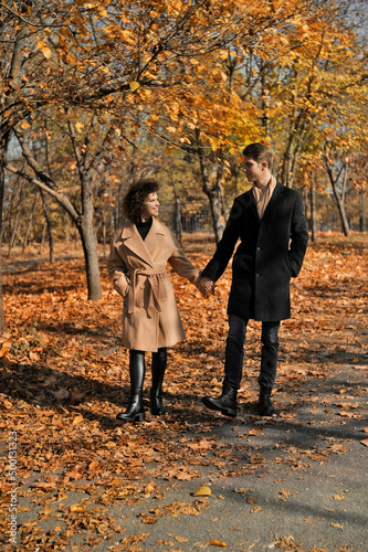 Young man in dark coat and woman in trendy beige coat with cup of coffee outdoor. Beautiful young couple drinks coffee in the autumn park with falling foliage.  © Alona