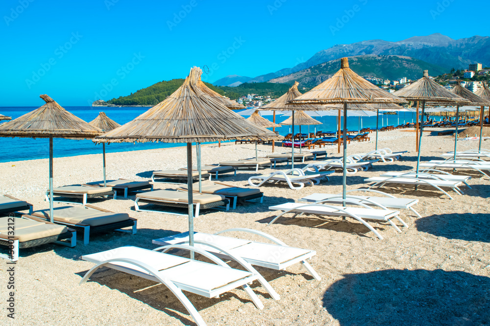 Straw beach umbrellas and comfortable sun loungers on clean sand and pebble beach. Himare. Albania.