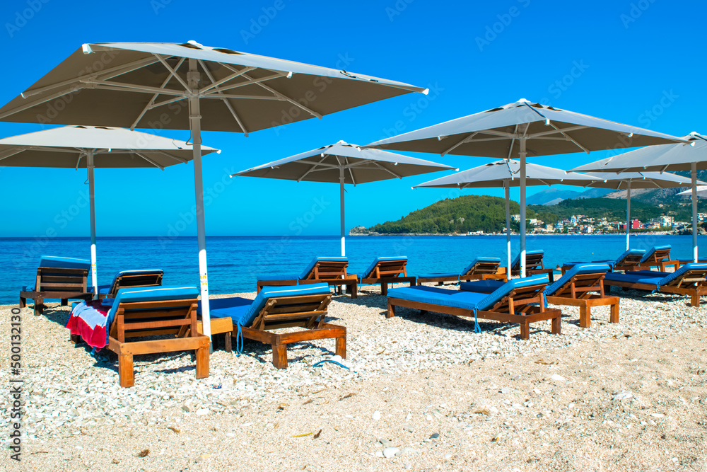 Grey beach umbrellas and comfortable sun loungers on clean sand and pebble beach. Himare. Albania.
