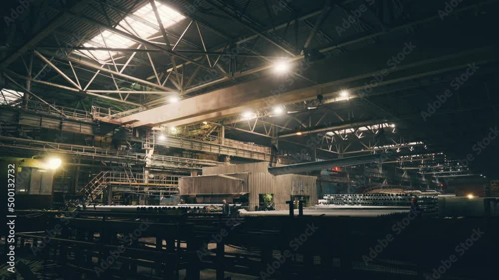 Large girder or beam crane transports iron pipes inside large dark workshop in metallurgical factory. Large girder or beam crane transports iron pipes inside large dark workshop in metallurgical factory.
