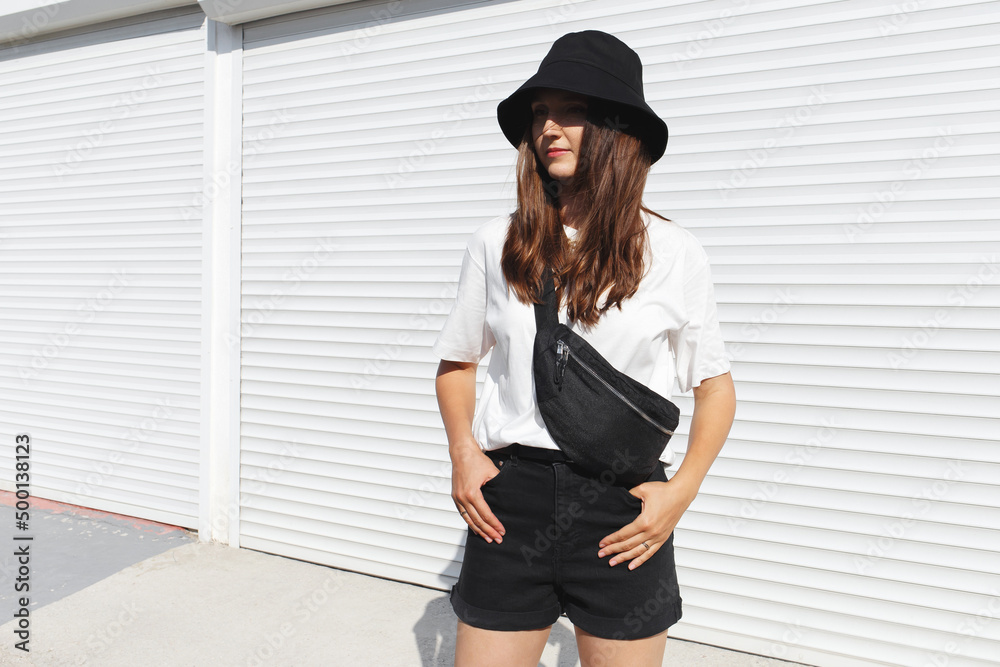 Woman, wearing white t-shirt, black shorts, fanny pack or waist pack and  bucket hat, standing outdoor near white wall. Details of stylish trendy  basic minimalistic casual outfit. Street fashion. Photos