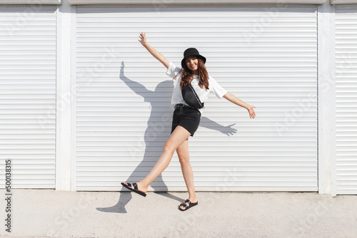 Woman, wearing white t-shirt, black shorts, fanny pack or waist pack, bucket hat and flat sandals, standing outdoor near white wall. Stylish trendy basic minimalistic casual outfit. Street fashion.