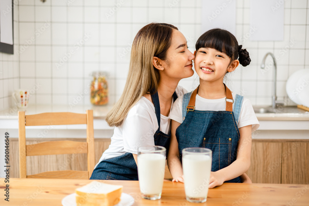 Asian teen girl drinking milk with her mom and eating bread, sitting at kitchen table.Loving mom kissing her little daughter in the kitchen, cheerful family .