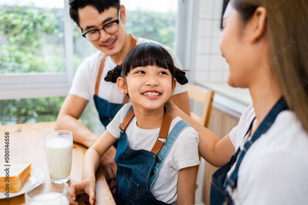 Fototapeta premium Asian family enjoying breakfast at cozy kitchen, little girl daughter sitting on table, drinking milk with smiling father and mother in morning. Happy family in kitchen.