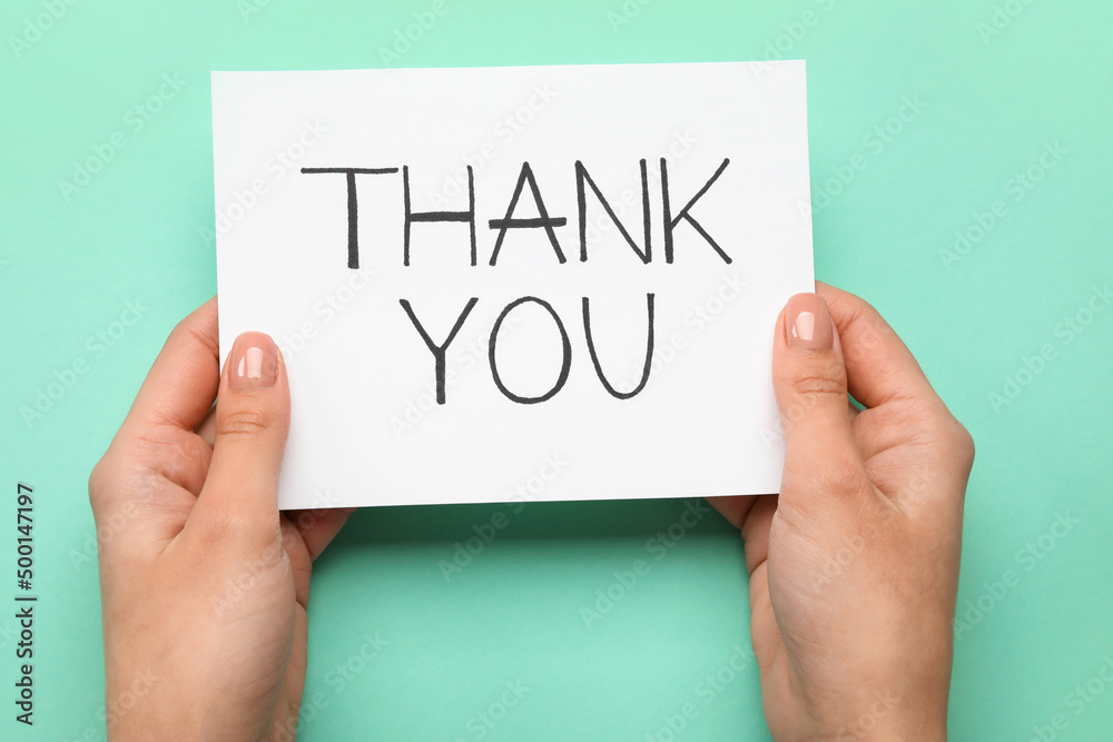 Female hands holding sheet of paper with text THANK YOU on turquoise background Female hands holding sheet of paper with text THANK YOU on turquoise background