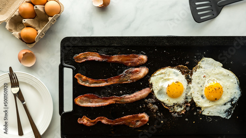 Fotografia Frying bacon and eggs on a cast iron griddle pan for breakfast.