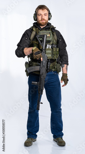 airsoft player in full growth. a man in an outfit, in headphones, a bulletproof vest, with a rifle and a pistol on a white background