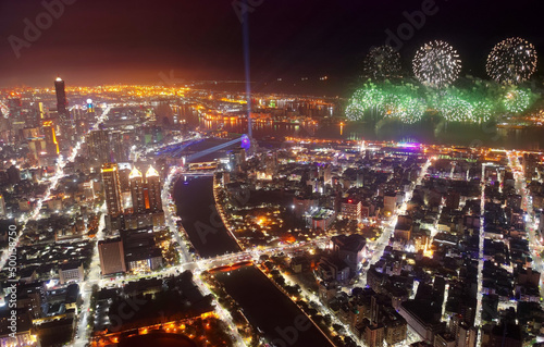 Aerial panorama of Kaohsiung City at dusk, a vibrant seaport in South Taiwan, with the landmark 85 Sky Tower standing among skyscrapers by the harbor & street lights dazzling with fireworks at dusk