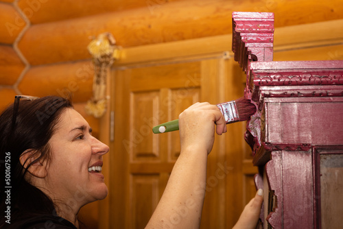 Woman with teeth smile on face painting ornamented cupboard in pink with big paint brush with doors in background. Reuse of old antique things. Home furniture renovation workroom.