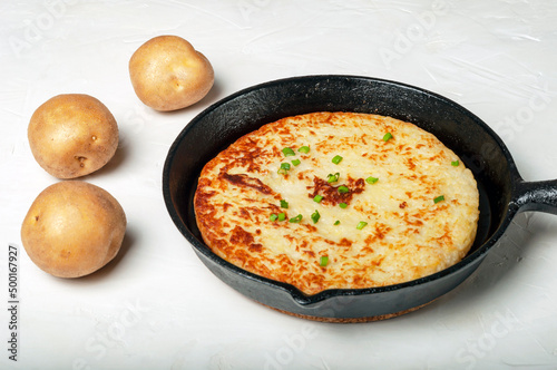 traditional Swiss dish is a potato pancake in a frying pan, decorated with green onions. whole tubers of unpeeled potatoes lie nearby. side view. photo