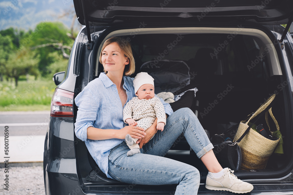 Mother with baby sitting in open car trunk outdoors. Road trip with baby. Travel in Europe. Young mother with her little baby having picnic near by the car. Safety driving by car with small kids. - obrazy, fototapety, plakaty 