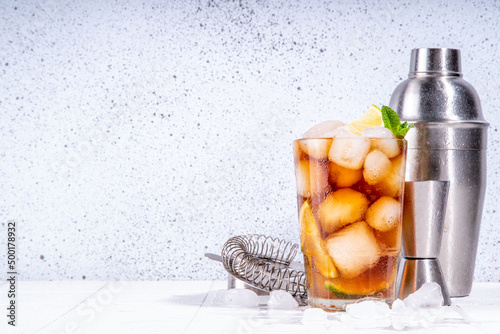 Rum, Lime and Cola Cuba Libre Cocktail. Summer cold Coca Coke drink with citrus juice, ice cubes and mint leaves, white tiled sun lighted background copy space