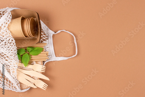 Kraft paper food cups and containers with wooden cutlery in white cotton net bag on light brown background with copy space - eco-friendly tableware. Sustainable food paper packaging concept