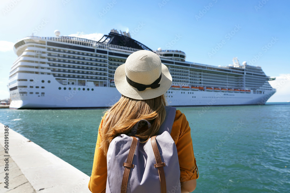 Cruise vacation. Rear view of tourist girl with backpack and hat standing in front of big cruise ship. - obrazy, fototapety, plakaty 