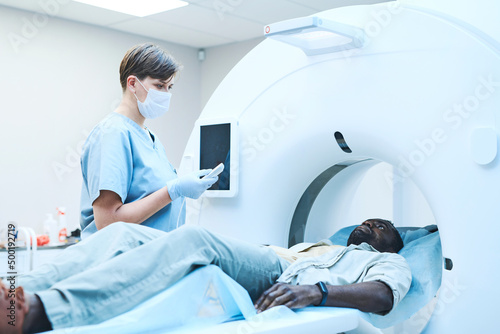 Young Caucasian MRI tech in mask pushing button on control panel while preparing patient for scanning photo