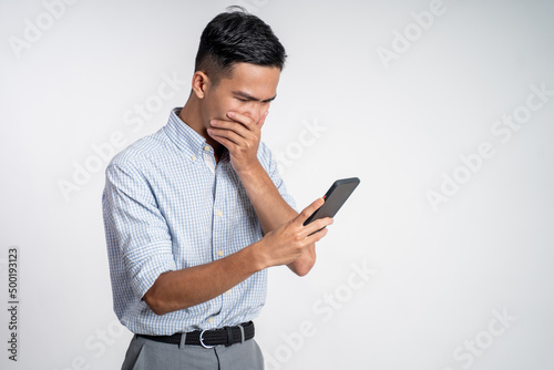 young asian man shocked while looking at smartphone screen on isolated background
