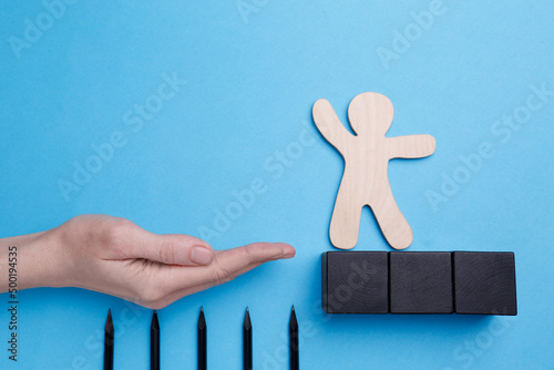 Woman holding hand to help human figure avoid trap with pencils as spikes on light blue background, closeup photo