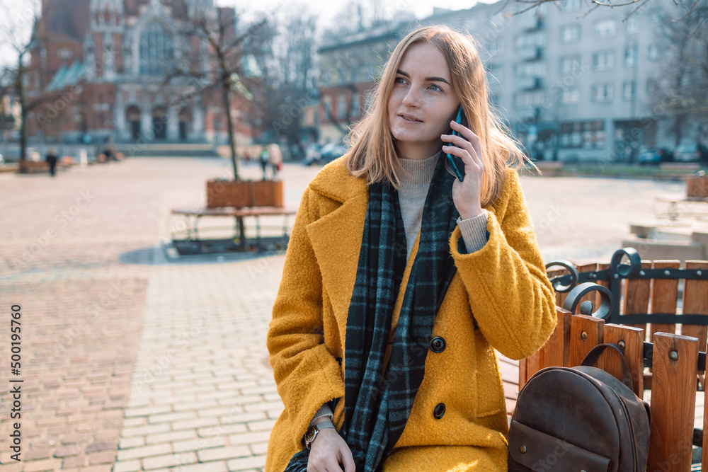 Fototapeta premium Beautiful smiling 30s woman with mobile phone walking on the street. Female calls by smart phone