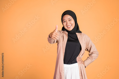 Asian woman in hijab smiling with thumbs up while standing on isolated background © Odua Images