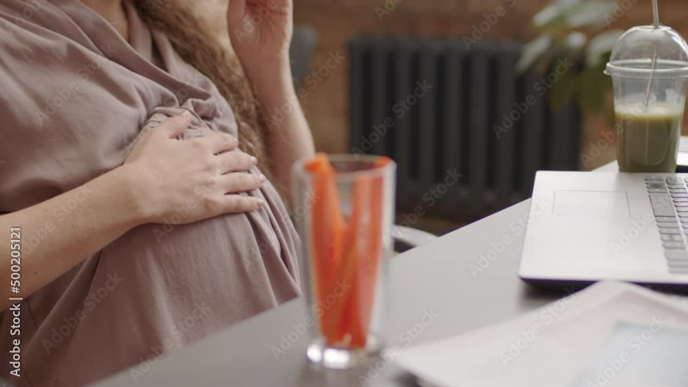 Midsection of cropped pregnant Caucasian woman sitting at desk in workplace at daytime, snacking with fresh sliced carrots, using laptop computer Midsection of cropped pregnant Caucasian woman sitting at desk in workplace at daytime, snacking with fresh sliced carrots, using laptop computer